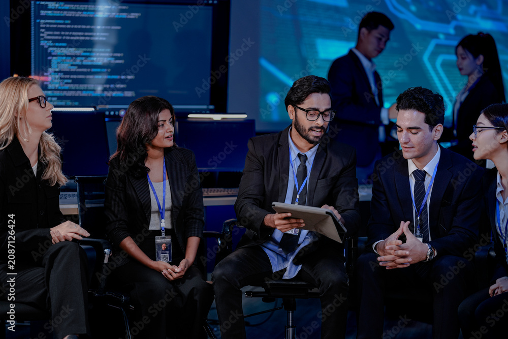 Obraz premium A large, diverse team of business professionals in a group meeting. An Indian man with a tablet leads the discussion with Asian and Caucasian colleagues in a high-tech office.
