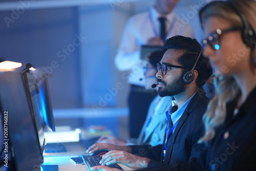 Foto Operators wearing headsets work at computers in a dark modern call center monitoring data on glowing screens at night