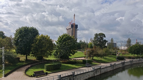A multi-storey concrete building is being erected on the banks of a river with concrete banks and in a city park with benches, stairs, lampposts and trees using tower cranes. Cloudy summer weather