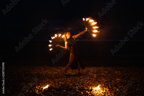 Female fire performer spinning flaming fans on a pebble beach at night.