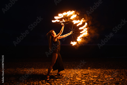 Female fire performer spinning flaming fans on a pebble beach at night.