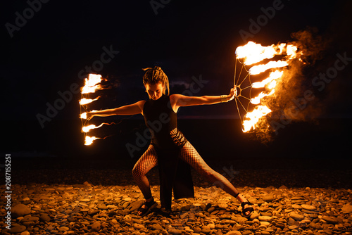 Female fire performer spinning flaming fans on a pebble beach at night.
