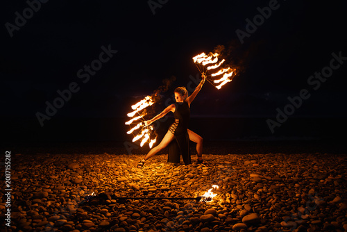 Female fire performer spinning flaming fans on a pebble beach at night.