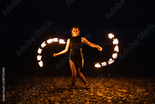 Female fire performer spinning flaming fans on a pebble beach at night.