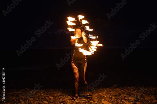 Female fire performer spinning flaming fans on a pebble beach at night.