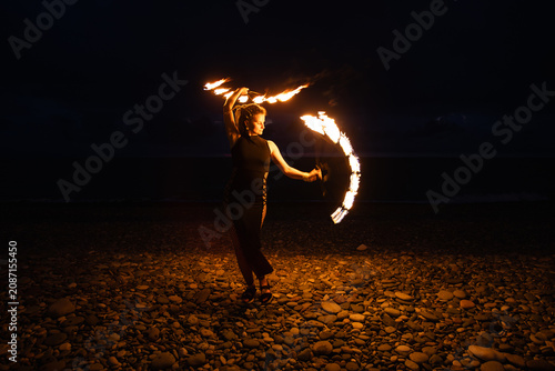 Female fire performer spinning flaming fans on a pebble beach at night.