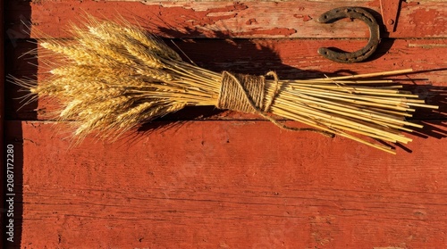 A rustic bundle of golden wheat stalks tied with twine resting on a weathered red wooden barn door with a metal horseshoe