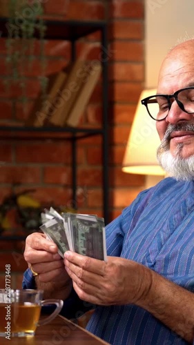 Indian senior retired man in kurta counting five hundred rupee notes while sitting at home on desk with computer, managing finances with focus