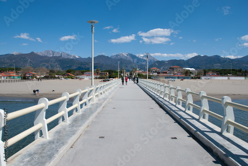 Modern pier leading to a sandy beach with dramatic snow-capped mountains under a clear blue sky, scenic coastal landscape, travel destination, peaceful outdoor panorama.