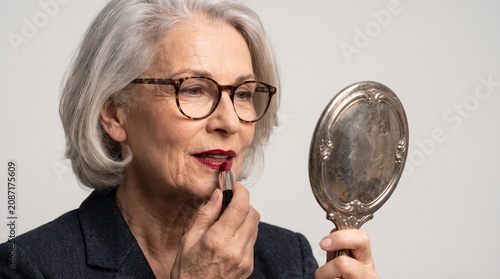 Senior woman with grey hair applying red lipstick using hand mirror