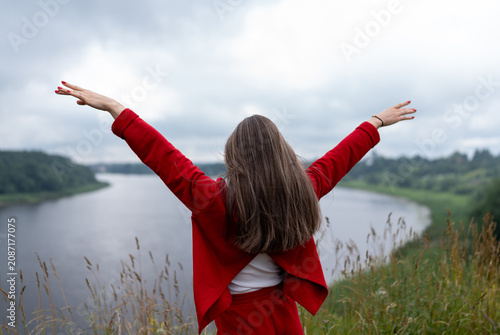 Fotografie Young woman in red outfit stands with open arms by a calm river, feeling freedom