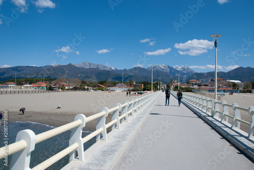 Modern pier leading to a sandy beach with dramatic snow-capped mountains under a clear blue sky, scenic coastal landscape, travel destination, peaceful outdoor panorama.
