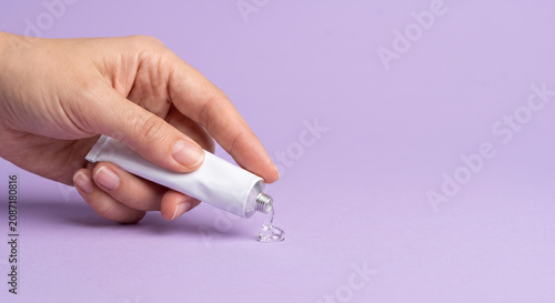 Close up of female hand squeezing transparent moisturizing gel out of white tube onto purple background.