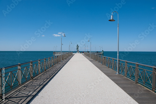 Modern pier in Versilia extending into the calm Mediterranean Sea under a clear blue sky, featuring a sculpture, symmetrical perspective, tranquil coastal travel destination.