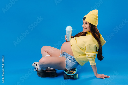Fotografie a pregnant girl in yellow clothes with a glass of juice sits on a skateboard on
