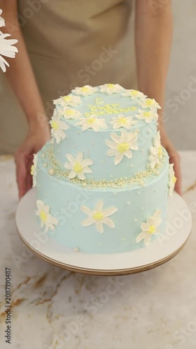 Pastry chef hands spinning a light blue two-tiered cake decorated with white chamomile flowers