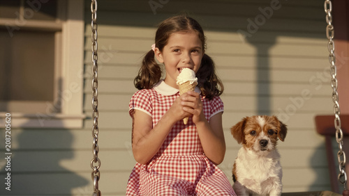 A happy young girl enjoys an ice cream cone with her cute dog on a sunny porch swing. This joyful childhood scene captures summer fun and friendship