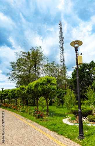 Parks and cell tower in a green space. People walk along a path in a park with trees and flowers under a cloudy sky. A cell tower stands in the background.