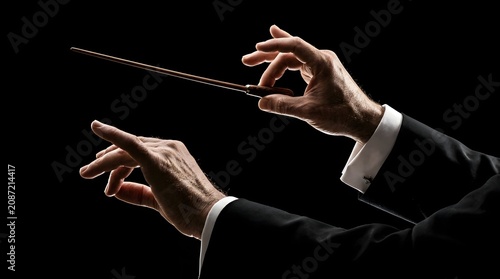 A detailed close-up of a professional orchestra conductor's hands, one holding a wooden baton, performing a dramatic musical movement against a stark, solid black background.
