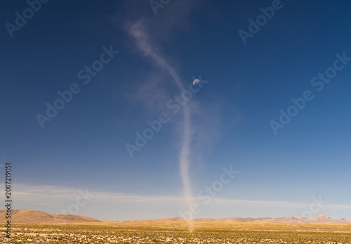 A tall dust devil whirls upward across the vast, arid Altiplano plains toward a visible daytime moon under a deep blue sky in Peru
