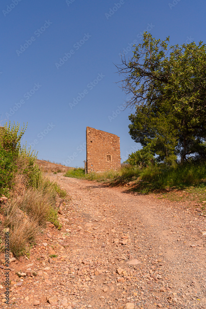 Naklejka premium Wanderlust meets history on a sunbaked gravel path leading to a crumbling medieval stone tower in Valencia Province. Perfect for travel, heritage, and adventure editorial use.