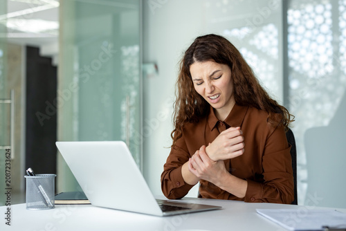 Businesswoman experiencing wrist pain or discomfort while working on her laptop at a modern office desk, an issue often stemming from repetitive strain injury or carpal tunnel syndrome