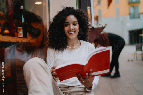 Beautiful young woman holding open red book and looking at camera with happy smile on cozy cafe terrace, female student studying outdoor, youth education and lifestyle concept portrait