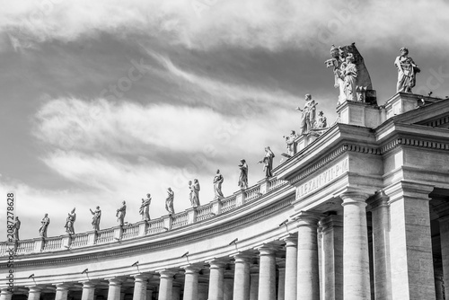 Curved classical colonnade with row of statues on entablature under dramatic cloudy sky, black and white architectural photograph