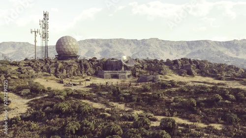 A remote landscape features a radar station surrounded by rocky terrain and greenery. Towering mountains rise majestically in the background, embraced by gentle clouds and a calm atmosphere.