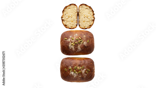 Two whole loaves of dark brown bread topped with pumpkin seeds and two slices of bread arranged vertically on a plain white background surface