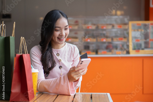A happy Asian woman sits at a table and uses her smartphone to make a credit card payment next to shopping bags.