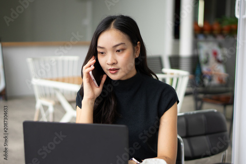 A focused Asian woman talks on her mobile phone while working on a laptop at a cafe.