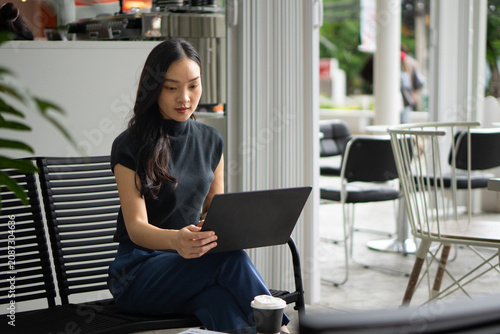 An Asian woman works comfortably on her laptop while sitting on an outdoor cafe bench next to a coffee cup.