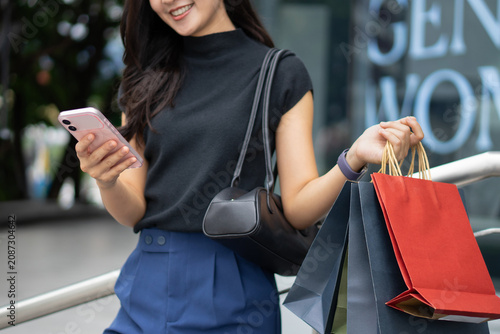 A close-up shot of a smiling Asian woman using her smartphone while holding shopping bags.