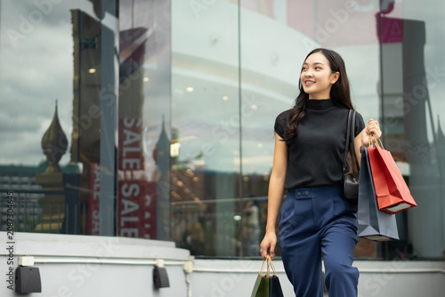 A cheerful Asian woman walks up outdoor steps holding colorful shopping bags in front of a modern glass building.