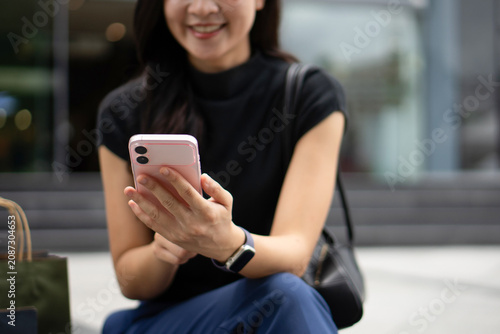 A close-up view focuses on an Asian woman's hands using a pink smartphone outdoors.