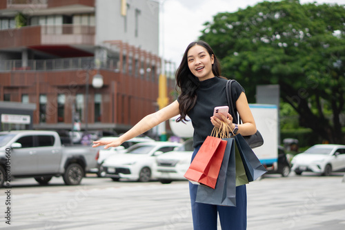 A cheerful Asian woman raises her arm to hail a taxi while holding shopping bags on a city street.
