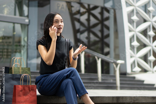 An expressive Asian woman talks on her mobile phone while sitting on outdoor concrete steps with her shopping bags.
