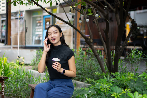 A cheerful Asian woman enjoys a phone conversation while holding a coffee cup on an outdoor bench surrounded by greenery.