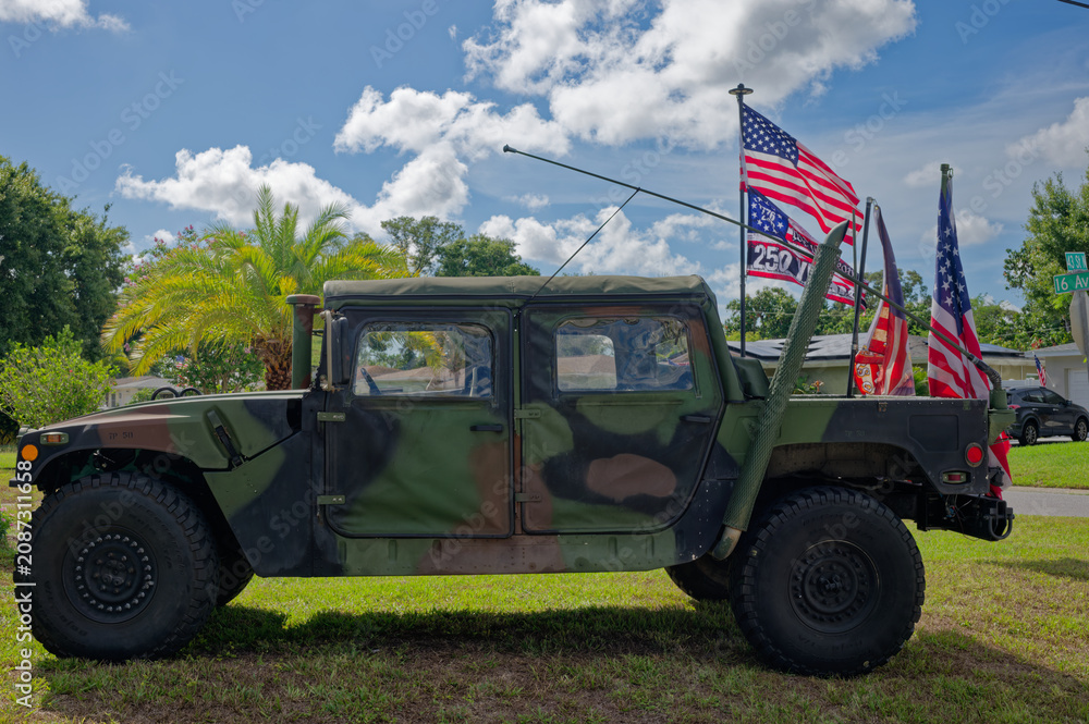 Fototapeta premium Camouflaged military Humvee (HMMWV) parked on a suburban lawn with several American flags and banners. Editorial Use Only July 5, 2026 St. Petersburg , FL USA. Evoking patriotism, veterans, and milita