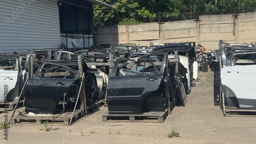 Rows of stripped car parts sit in a scrapyard. Metal doors and frames stand on wooden pallets. Sunlight highlights decayed paint and rust. Perfect for automotive salvage, recycling