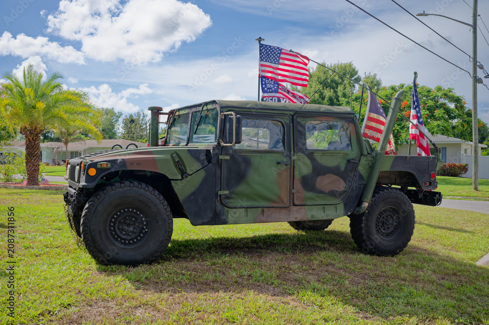 Fototapeta premium Camouflaged military Humvee (HMMWV) parked on a suburban lawn with several American flags and banners. Editorial Use Only July 5, 2026 St. Petersburg , FL USA. Evoking patriotism, veterans, and milita