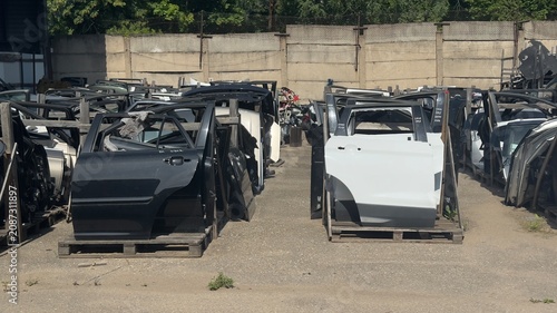 Rows of stripped car parts sit in a scrapyard. Metal doors and frames stand on wooden pallets. Sunlight highlights decayed paint and rust. Perfect for automotive salvage, recycling