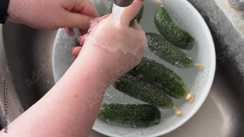 A close-up shot of hands thoroughly washing freshly harvested green cucumbers in a bowl under running tap water, focusing on food hygiene, preparation, and healthy organic lifestyle.
