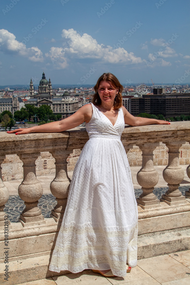 Fototapeta premium Smiling young woman in white summer dress poses on stone balcony overlooking historic cityscape on bright sunny day during her beautiful European summer vacation travels
