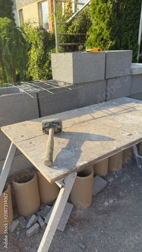 Concrete blocks stacked for construction. Hammer rests on work surface. Poured concrete forms base. Metal supports hold structure upright. Outdoor building site under sun. Ideal for construction, DIY