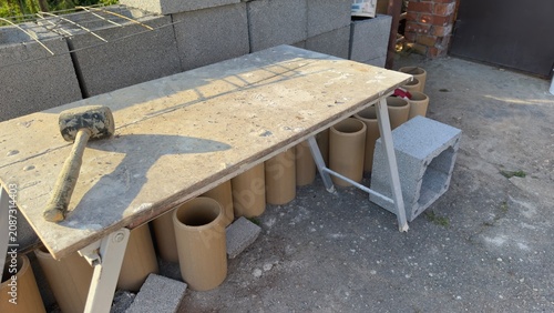 Concrete blocks stacked for construction. Hammer rests on work surface. Poured concrete forms base. Metal supports hold structure upright. Outdoor building site under sun. Ideal for construction, DIY