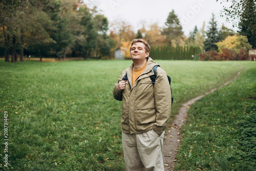Happy 30s blonde man with backpack standing in the countryside. Traveler. Autumn time. Positive handsome blonde male standing outdoor