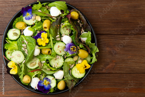 Colorful salad with greens cucumbers olives cheese and pansies flowers on a wooden table