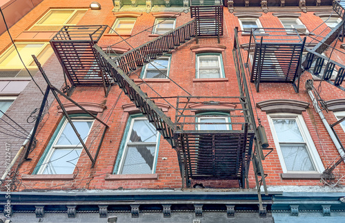 An eye-level view of a classic 3-story residential walk-up building in Manhattan, New York City. The traditional urban architecture features an external fire escape.